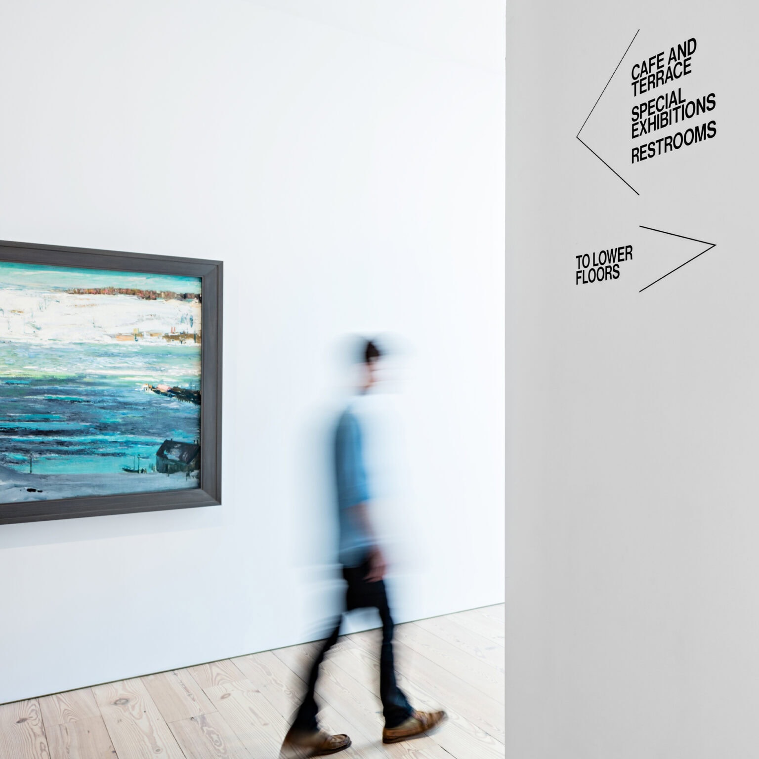 Whitney Museum minimal directional signage adhered to wall of white gallery. Painting appears in the background.