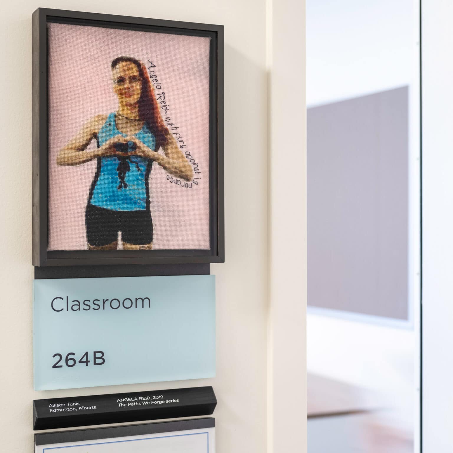 Framed Cross-stitch depicting a woman mounted to a room sign. Artist Credit: Allison Tunis,